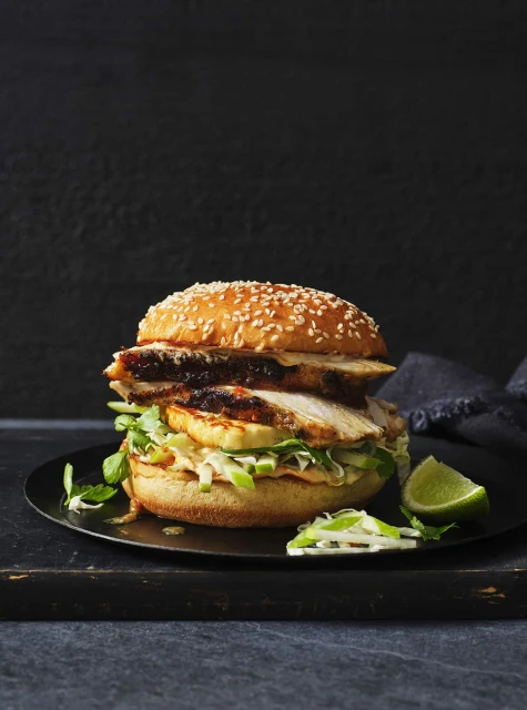 Sesame bun filled with grilled chicken, sauce, lettuce, lime wedge. Dark background with shallow depth of field creates a culinary aesthetic.