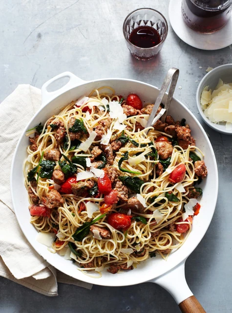 Overhead shot of spaghetti with sausage, spinach, and tomatoes served in a white pan on a gray surface with a linen napkin.