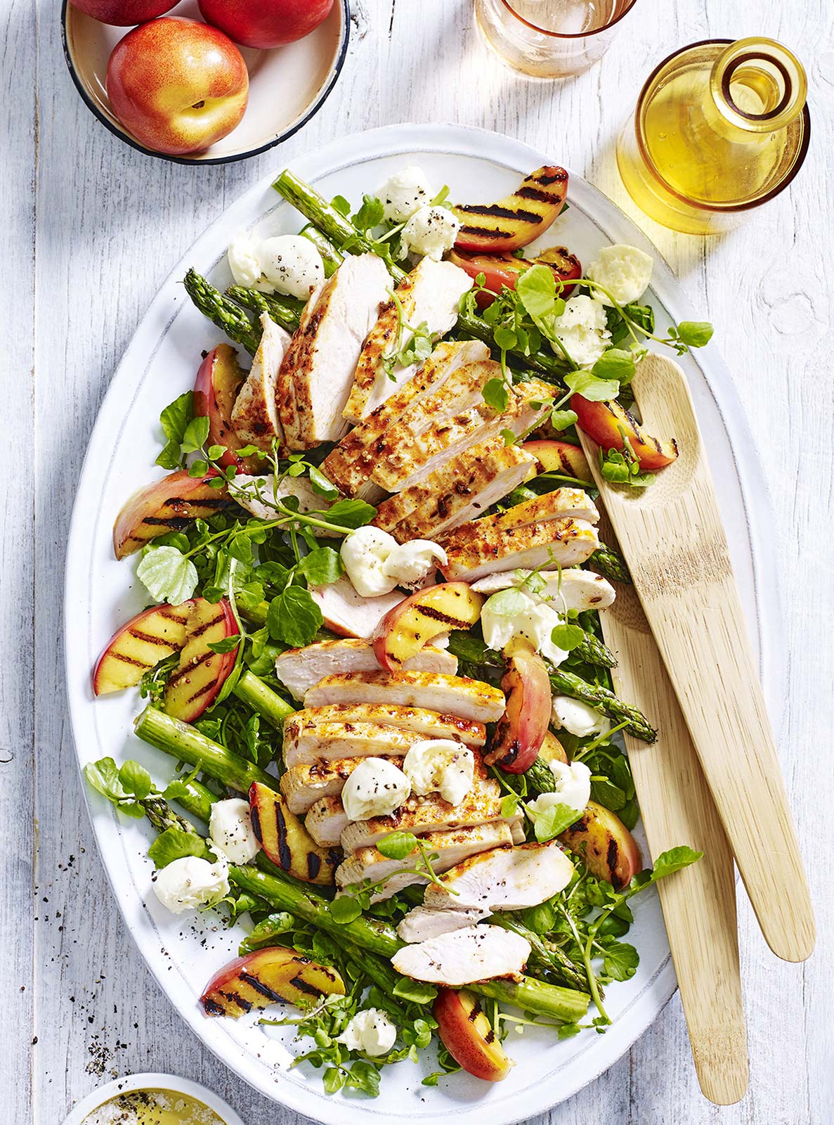 Overhead shot of a grilled chicken salad featuring peaches, asparagus, and watercress on a white platter with wooden serving utensils.