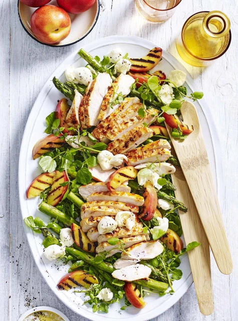 Overhead shot of a grilled chicken salad featuring peaches, asparagus, and watercress on a white platter with wooden serving utensils.