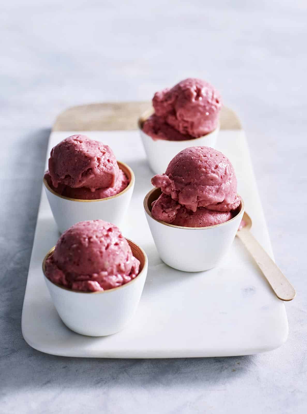 Four white bowls of reddish-pink ice cream on a marble surface with a wooden spoon on a bright background, food photography.
