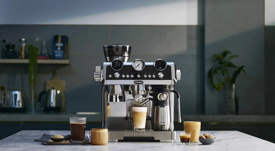 De'Longhi espresso machine on a marble countertop with glasses of coffee, a bowl of sugar, and mugs on a kitchen counter.