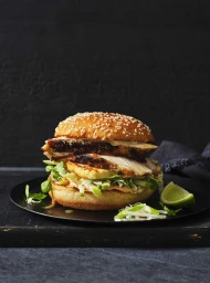 Gourmet chicken burger with sesame bun, sliced chicken, and herbs served on a black plate with a lime wedge