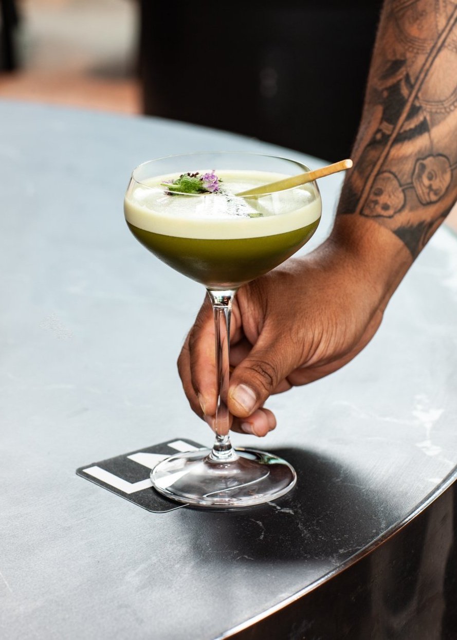 Close-up of a tattooed hand holding a cocktail in a stemmed glass garnished with flowers, set on a black table.