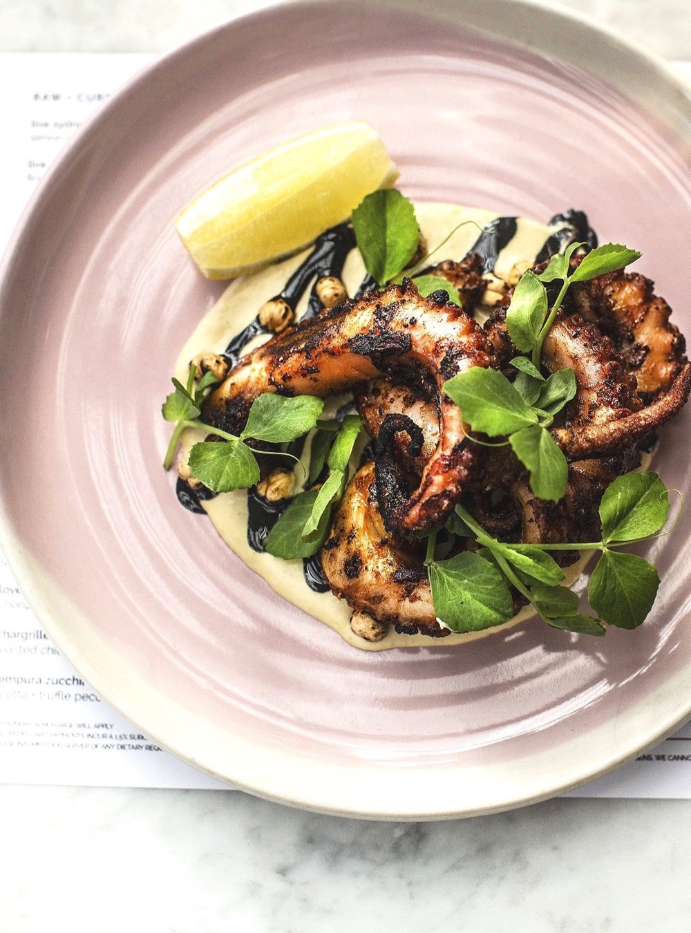 Overhead shot of grilled octopus appetizer on a pink plate, with lemon, herbs, and sauce, staged on a white marble table.