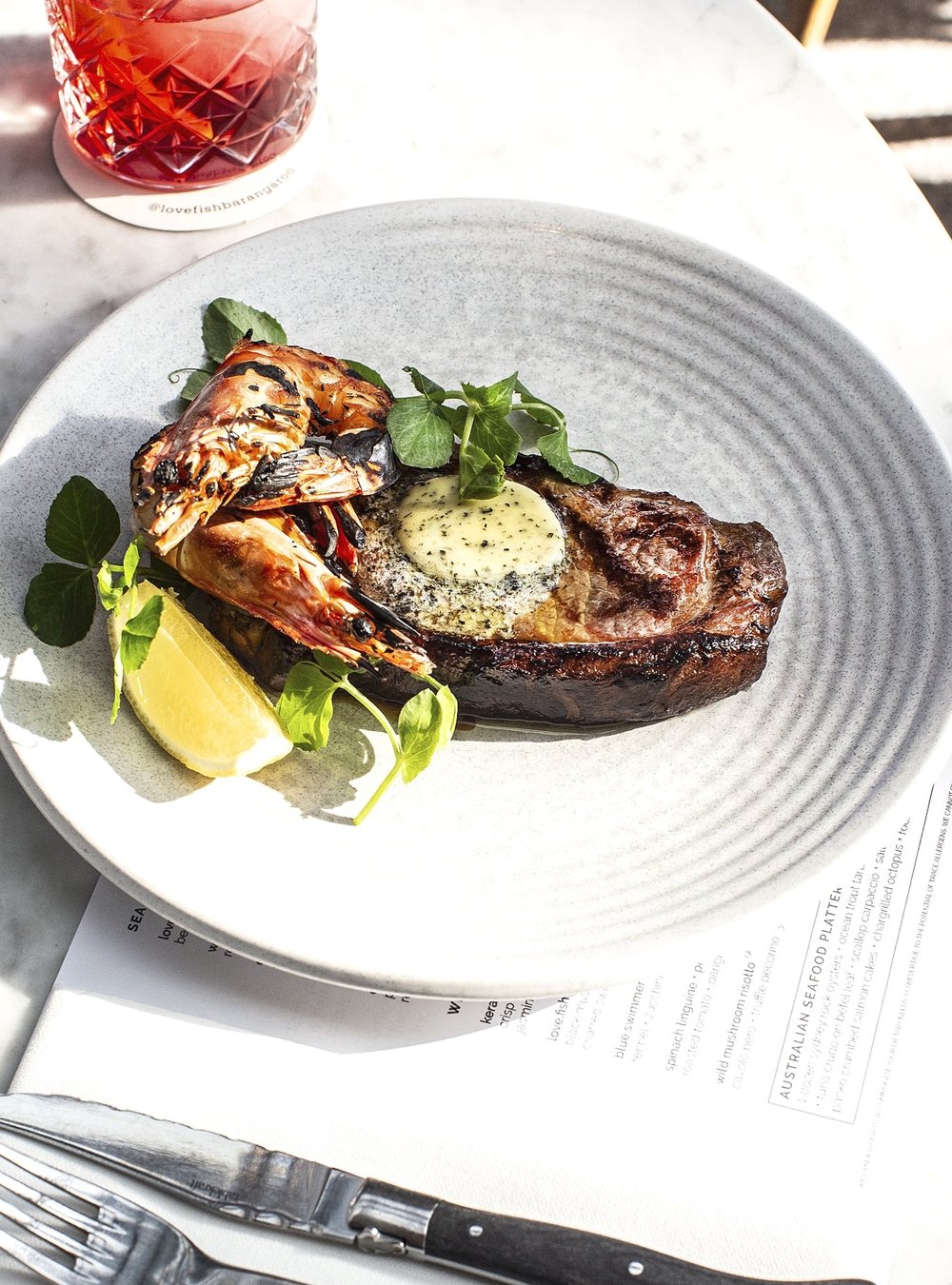 Overhead shot captures a beautifully plated steak topped with herb butter and grilled shrimp, served with a slice of lemon.