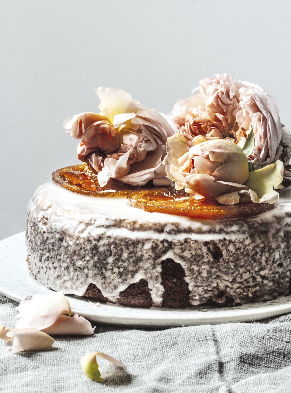 Close-up of a glazed cake decorated with fresh roses and candied oranges, sitting on a plate atop a linen tablecloth.