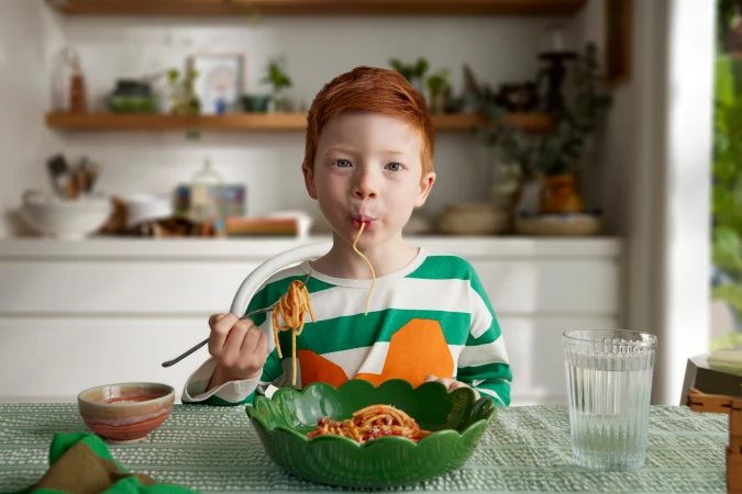 Red-haired child enjoys a plate of spaghetti at the table, wearing a striped shirt and sitting in a bright, naturally lit kitchen.