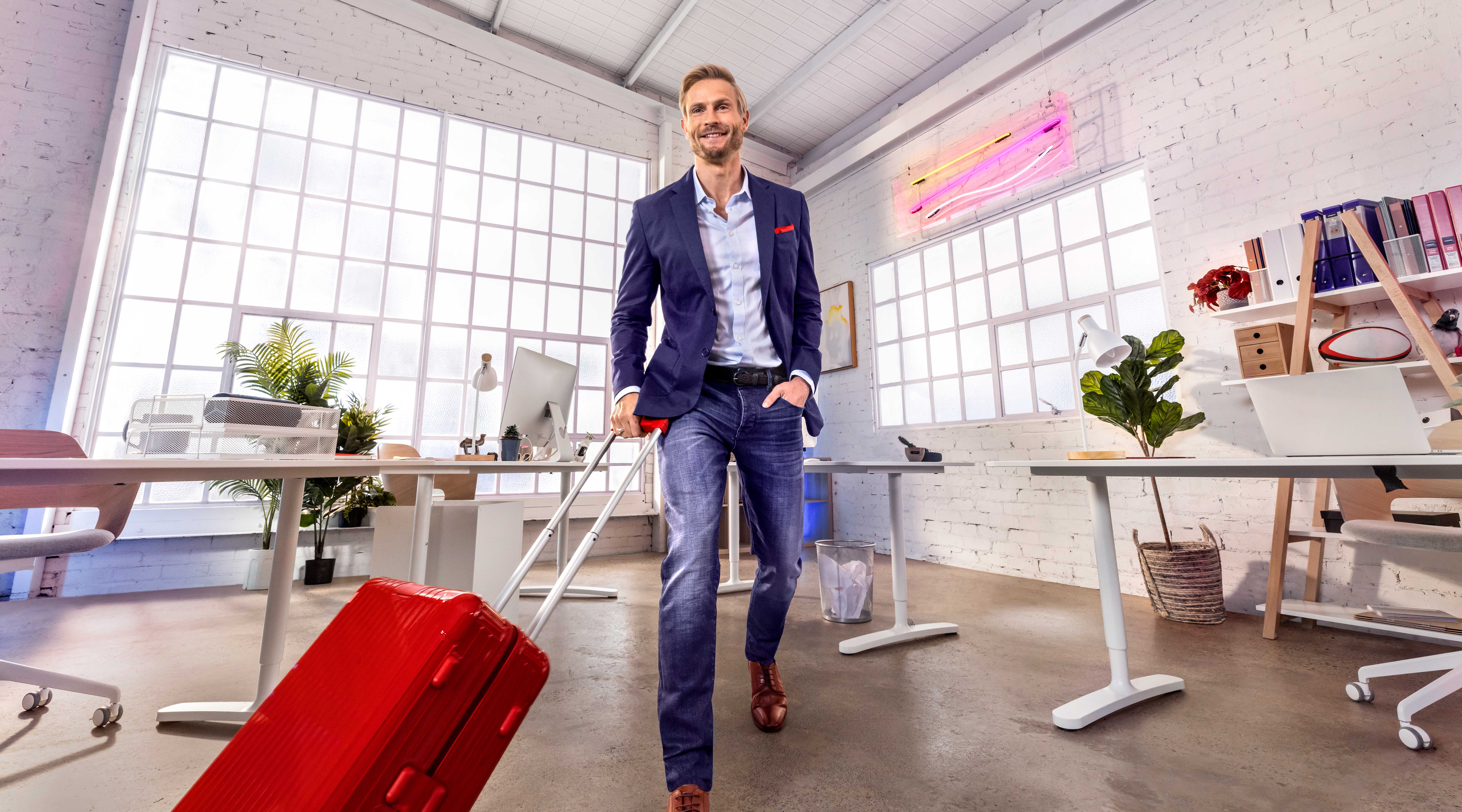 Smiling man in a blue suit pulling a red rolling suitcase in a modern office with white walls, large windows, and white desks.