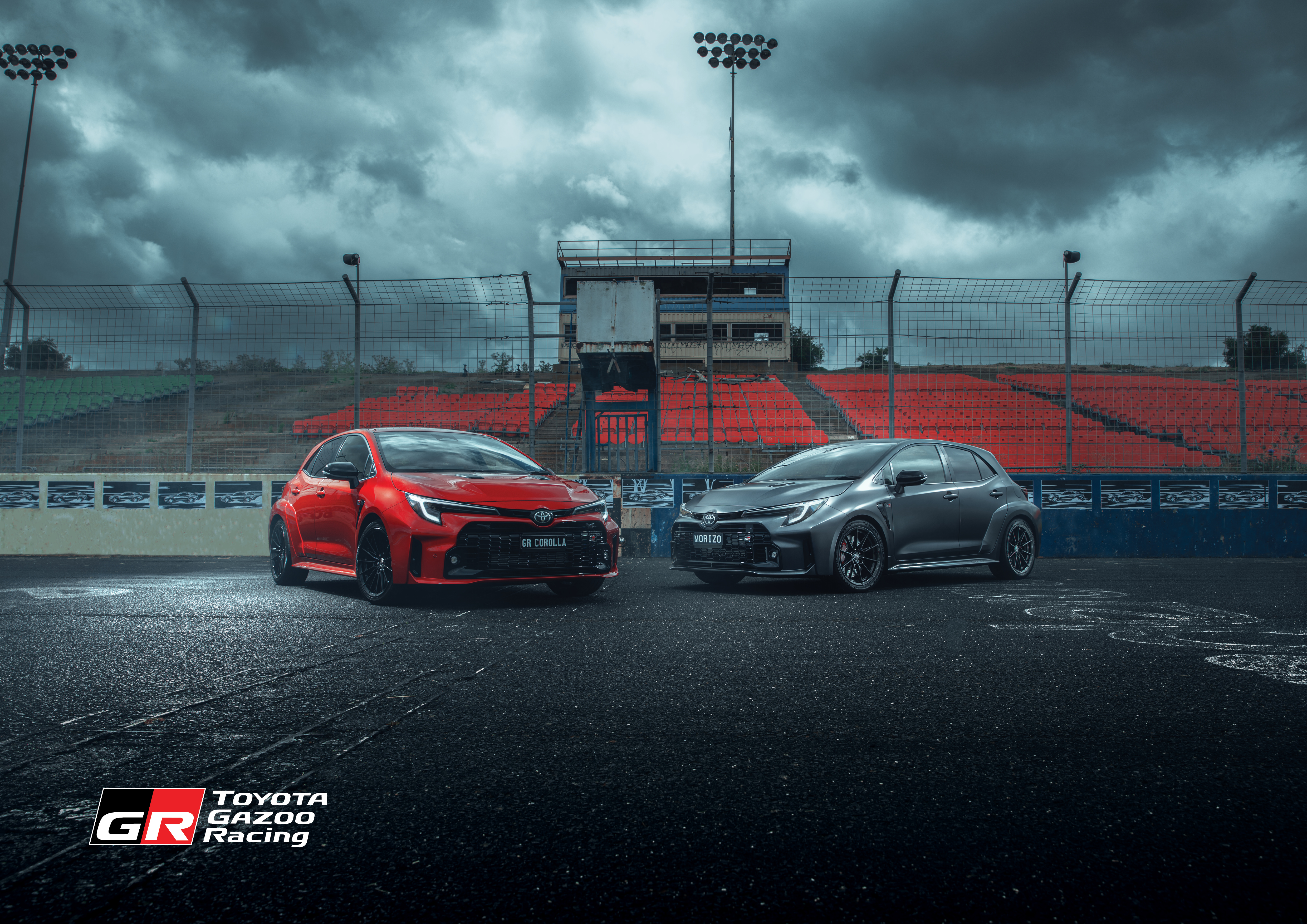 Two Toyota GR Corolla cars, one red and one gray, are parked on an asphalt track with grandstand seating in the background on an overcast day.