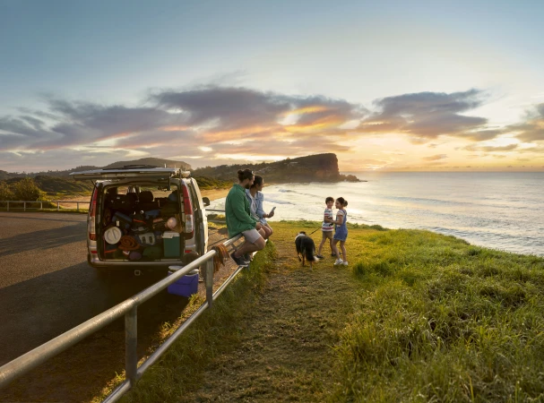 Family enjoys ocean view near their car packed with camping gear at sunset. Dog stands nearby. Travel, vacation, recreation.
