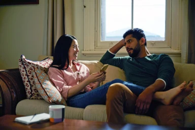 An Asian woman and a South Asian man relaxing together on a living room sofa, with natural light coming from a window behind them.
