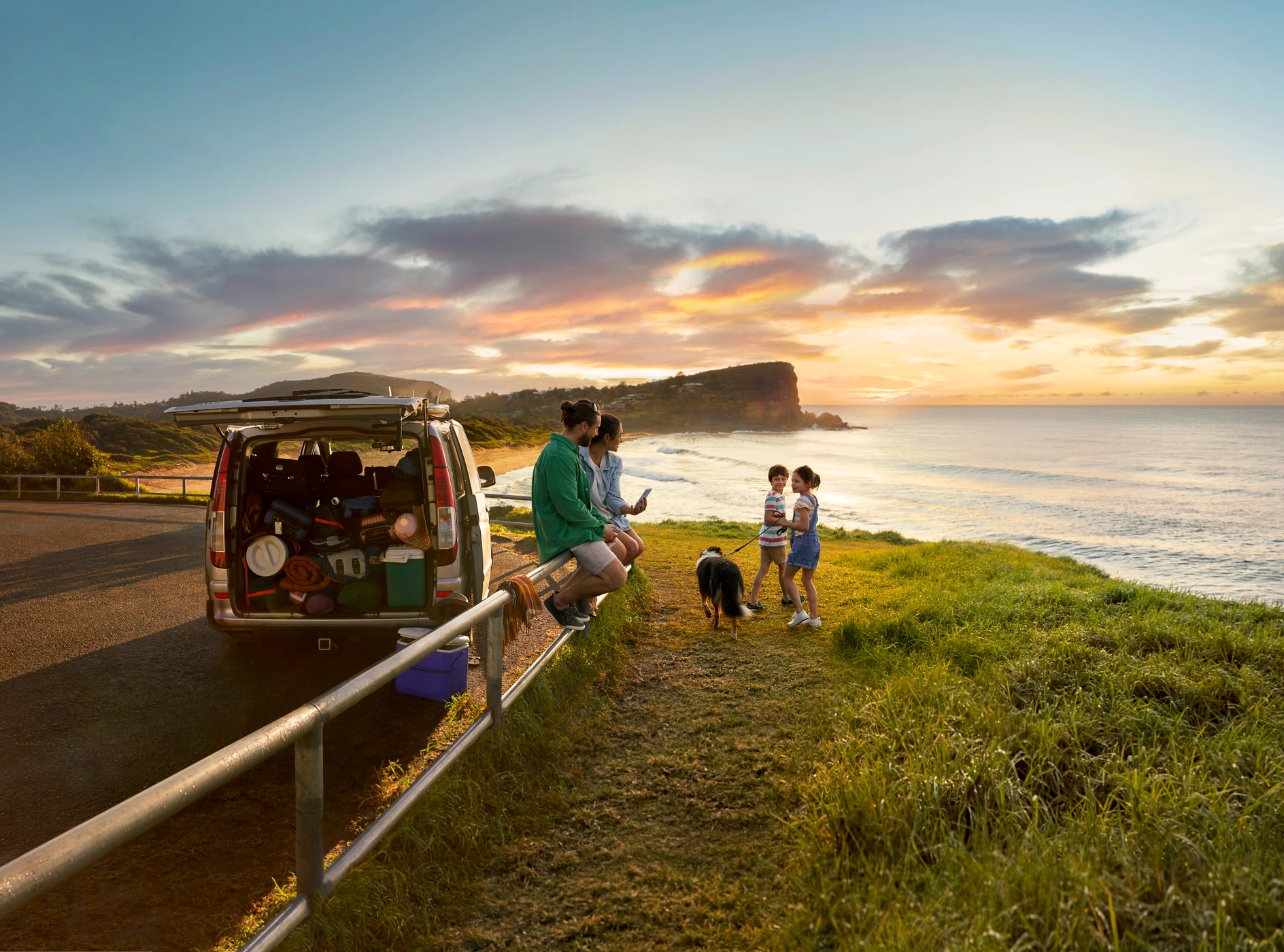 Family and dog enjoying sunset on a coastal clifftop next to a packed camper van with open rear doors, photographed in Sydney by Natalie Loveridge