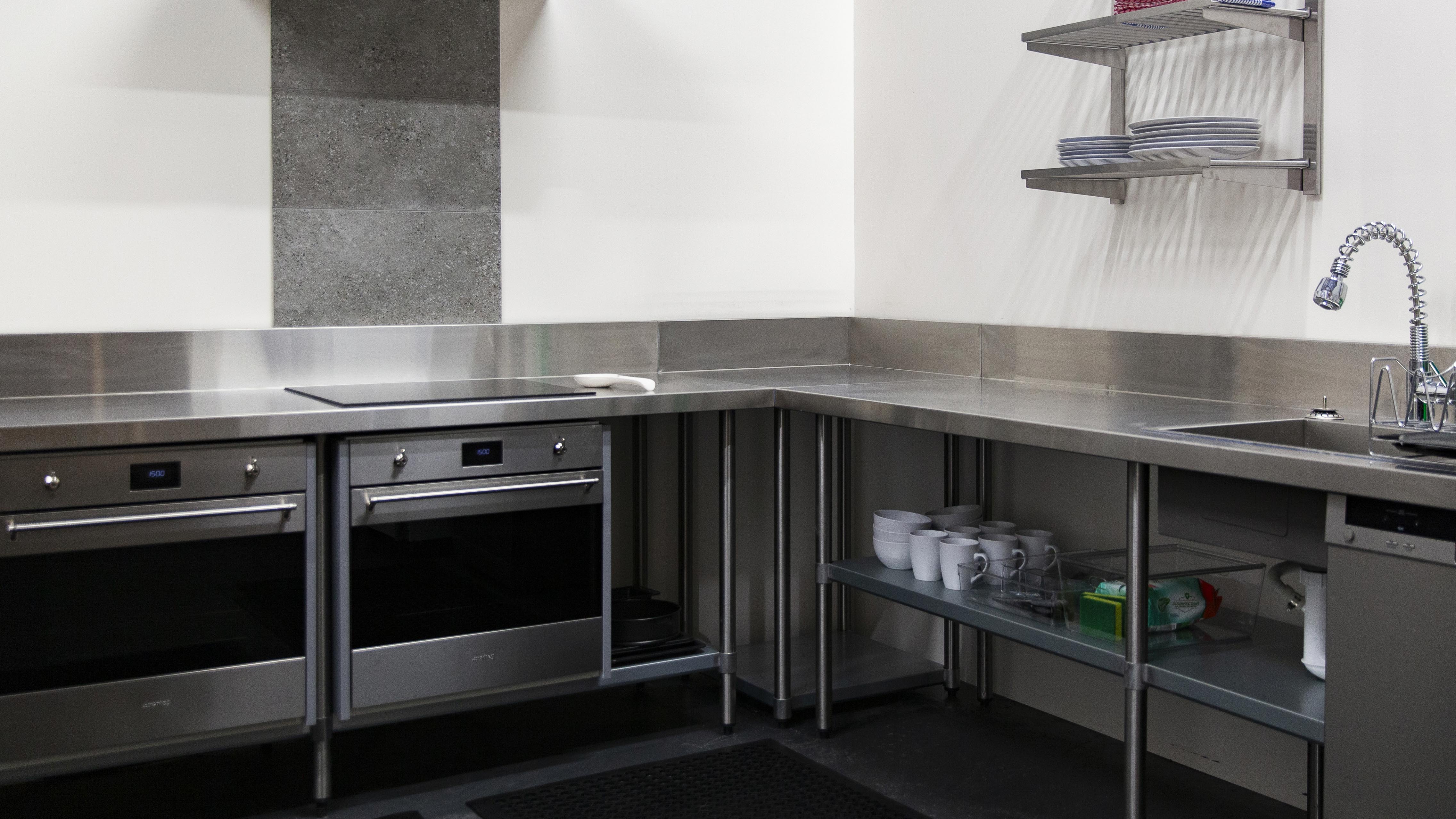 Stainless steel commercial kitchen with ovens, cooktop, shelving, sink, faucet, countertops and white walls with a grey tile accent.