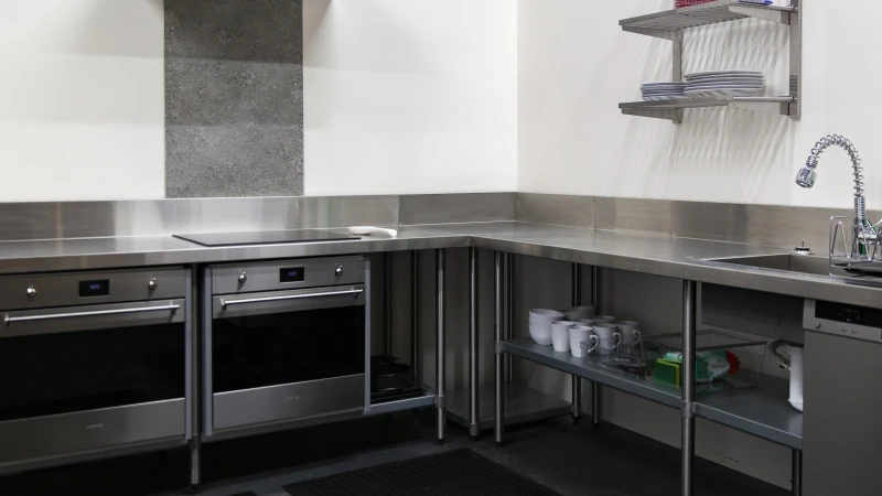 Stainless steel commercial kitchen with ovens, cooktop, shelving, sink, faucet, countertops and white walls with a grey tile accent.