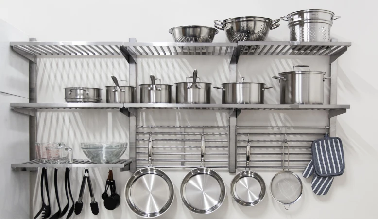 Stainless steel cookware and utensils on a metal shelving unit against a white wall in a home kitchen, studio, or showroom.