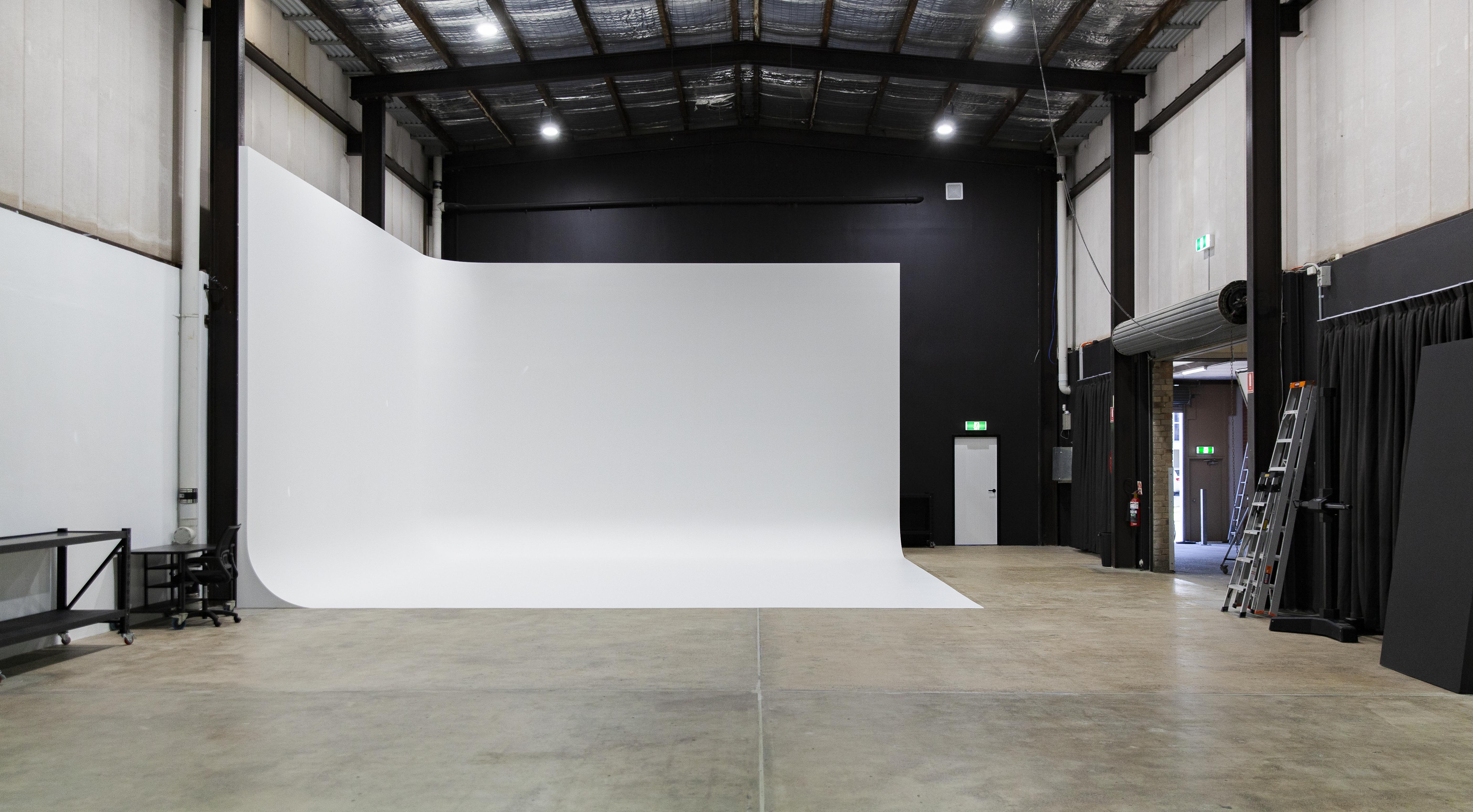 Wide photo studio with a curved white backdrop, industrial metal ceiling beams, concrete floor, and a ladder near an open doorway.