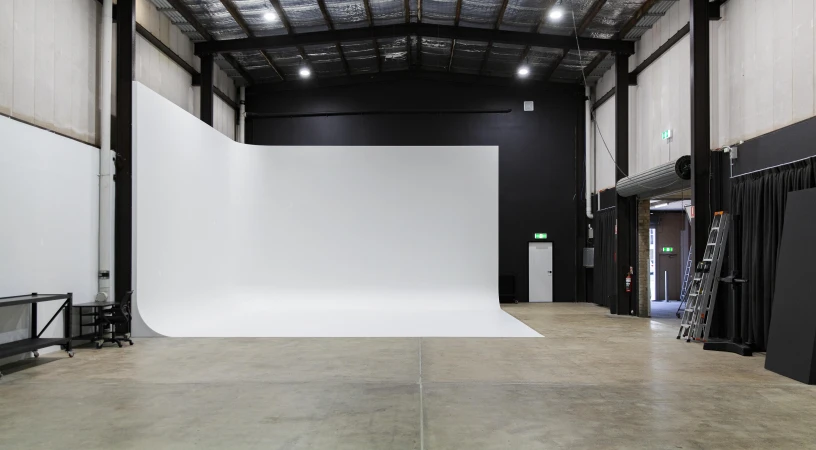 Wide photo studio with a curved white backdrop, industrial metal ceiling beams, concrete floor, and a ladder near an open doorway.