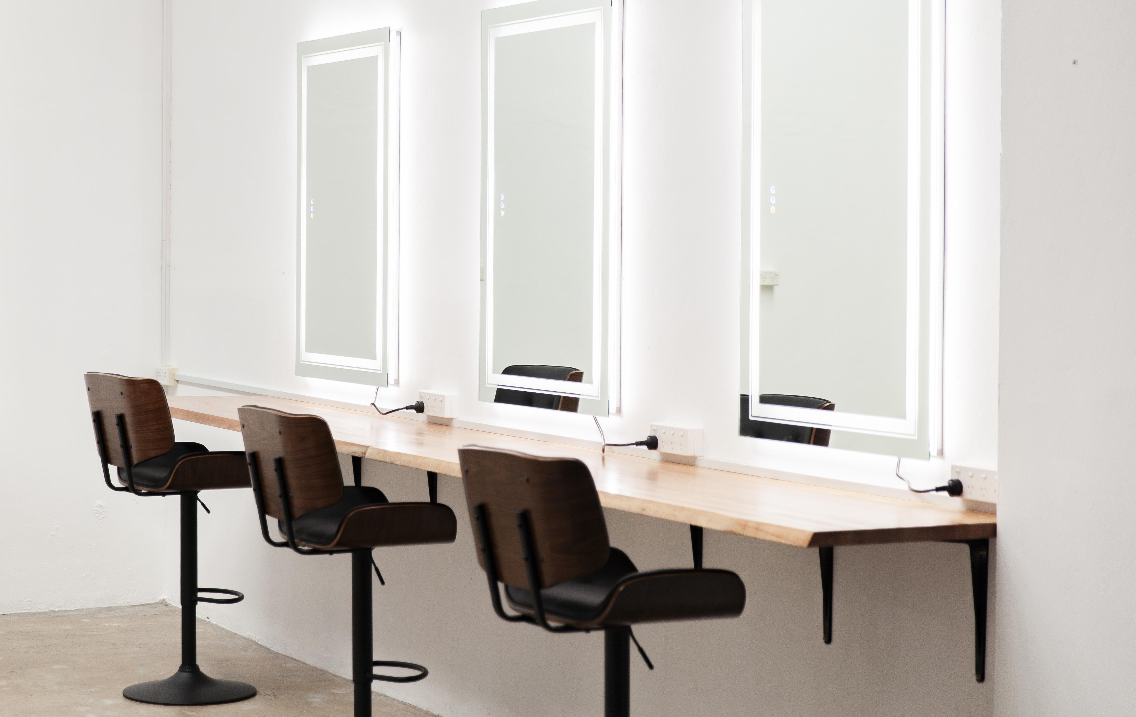 Photo of three wooden styling chairs facing lighted mirrors in a hair salon or studio space with white walls and a light wood counter.