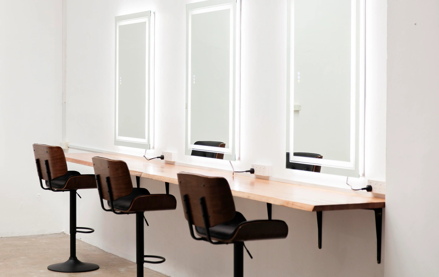 Three modern salon styling chairs with dark seats and wooden backs aligned along a long wooden counter with lighted mirrors.
