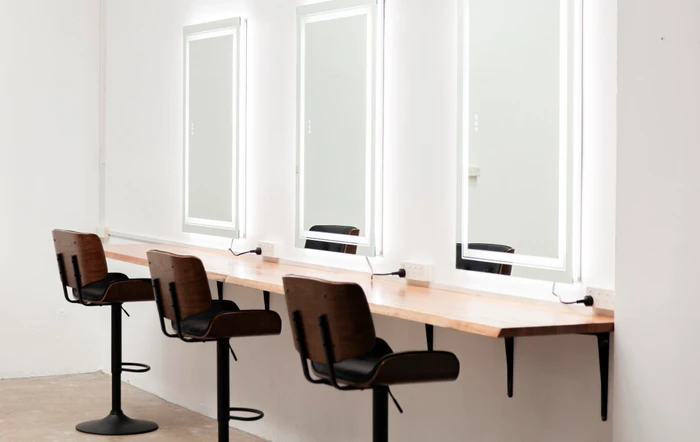 Three modern salon styling chairs with dark seats and wooden backs aligned along a long wooden counter with lighted mirrors.