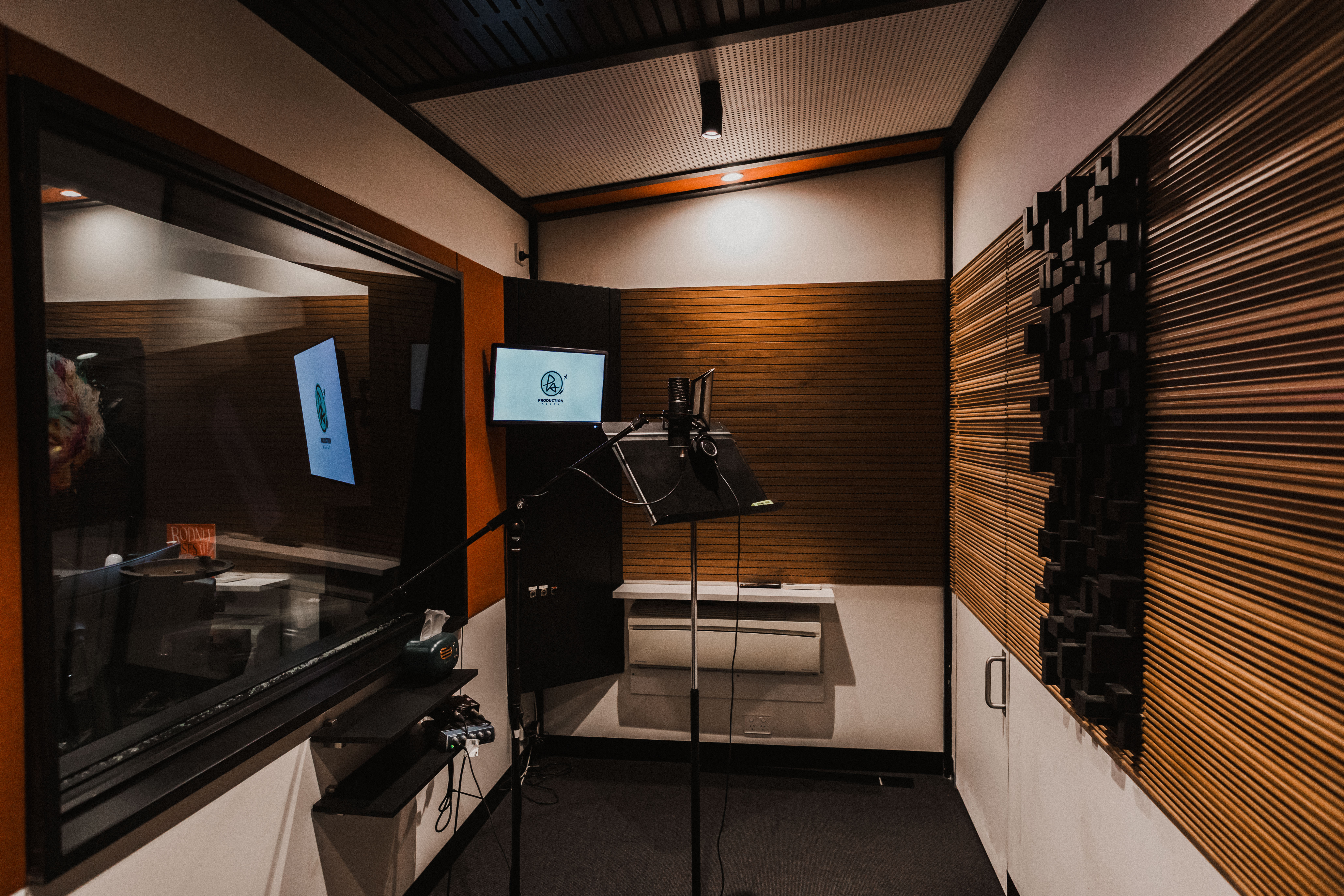 A professional microphone stands in a compact, soundproof recording booth with wood paneling and modern acoustic treatment.
