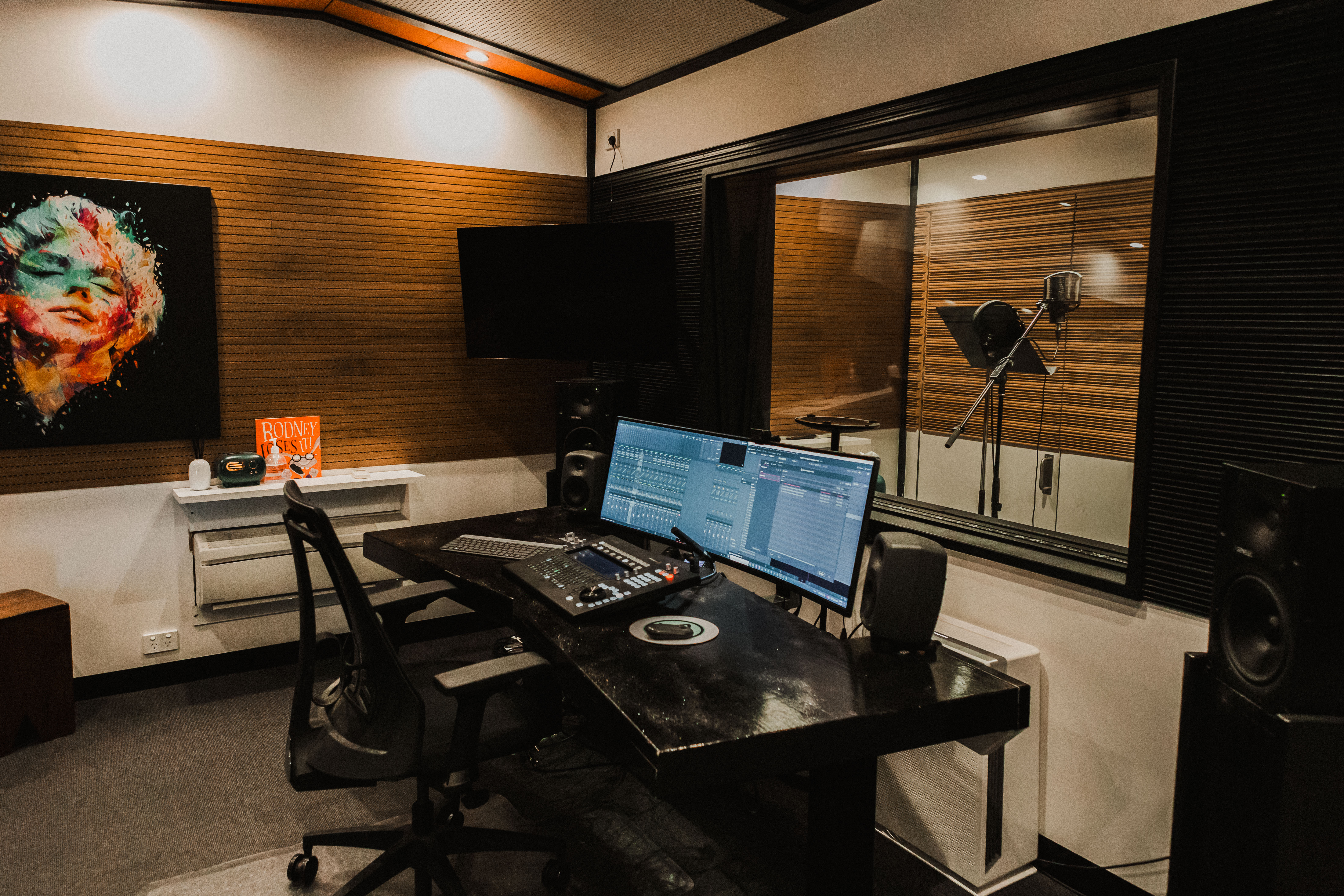 Interior view of a professional music recording studio with audio monitors, sound mixing equipment, microphone in sound booth, and artwork.