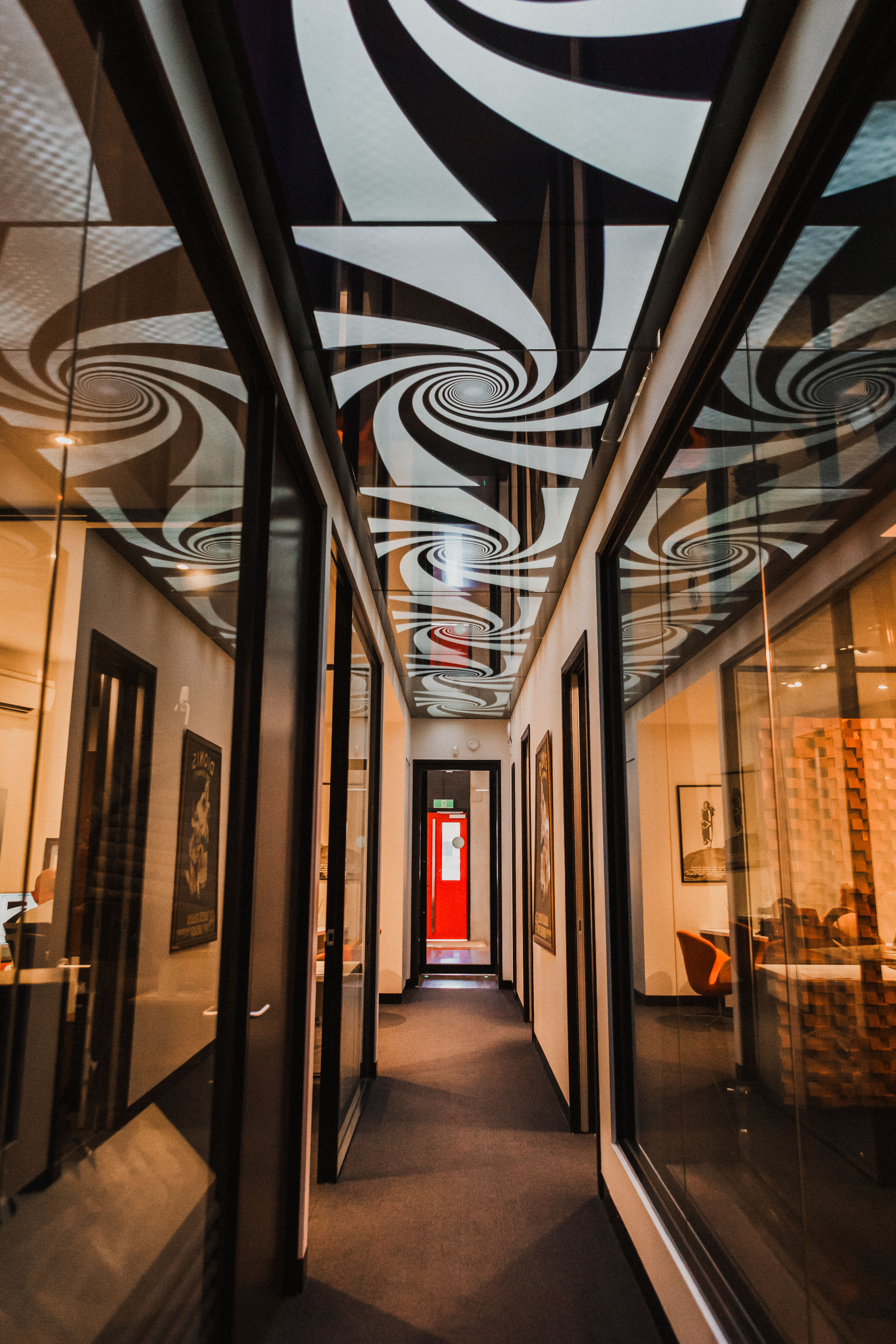 Interior shot of a long office hallway with a spiral pattern on the ceiling, doors to the offices along both sides and a red door in the distance.