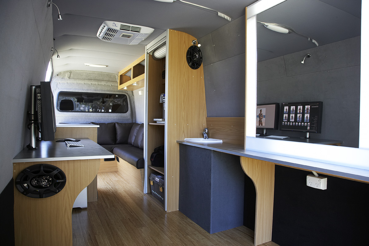 Interior view of a mobile studio van featuring a workstation with computer monitors, sink, cabinets, and seating area.