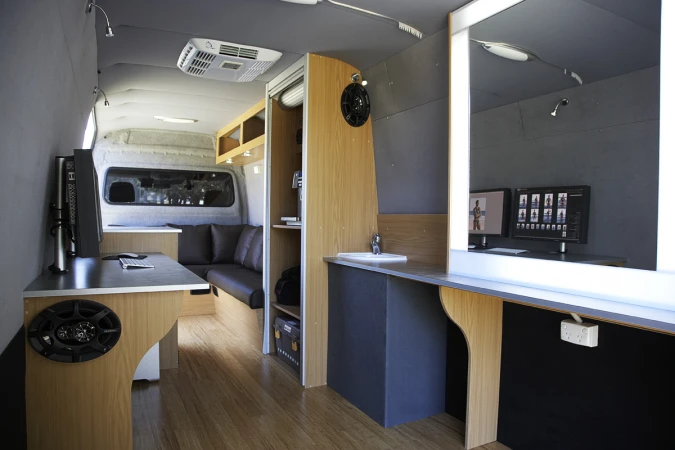 Interior view of a mobile studio van featuring a workstation with computer monitors, sink, cabinets, and seating area.