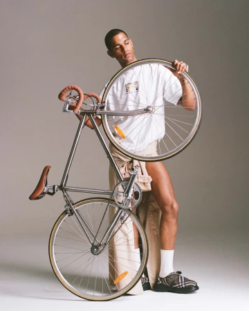 Young adult man poses indoors with a fixed gear bicycle. The bicycle is silver with a brown leather saddle and handlebar grips.
