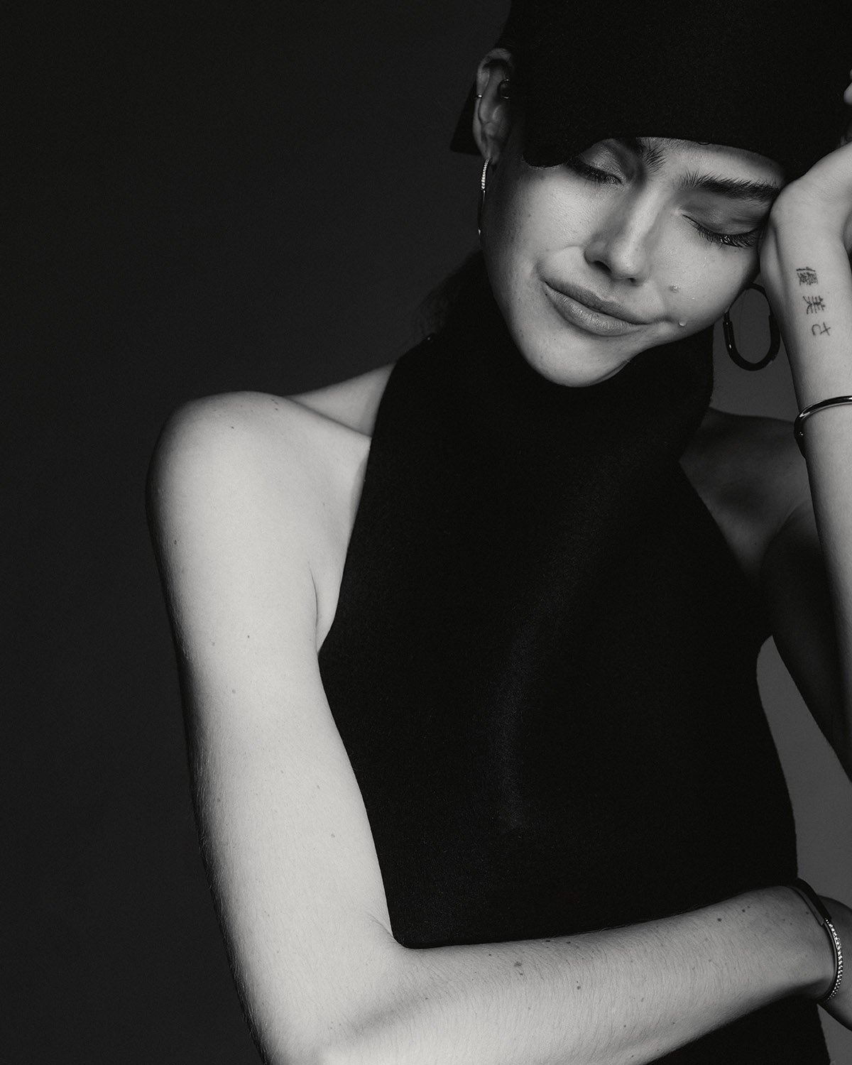 Monochrome portrait of a woman with dark hair, wearing a black halter top and cap, with a slight frown and jewelry.