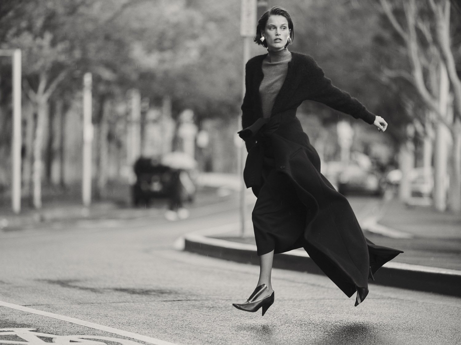 Black and white fashion shot of a woman in a long coat crossing a street. She is airborne against a blurred urban background.