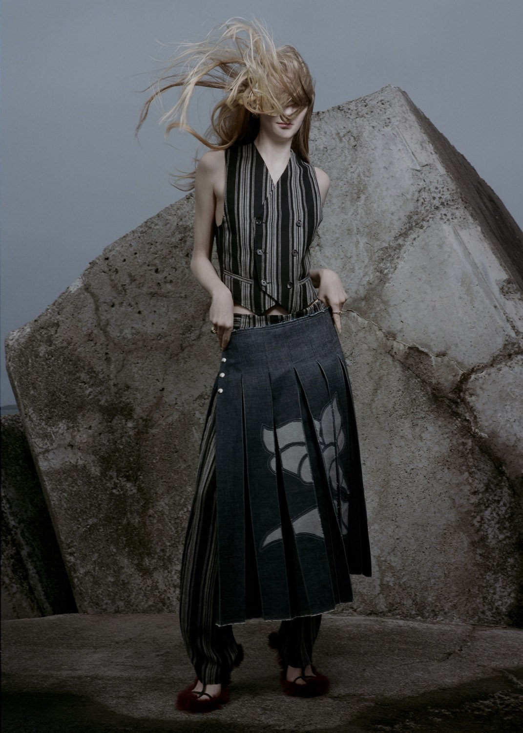 Fashion editorial shot of an adult woman in a striped vest and denim skirt posing near a large geometric concrete structure outdoors.