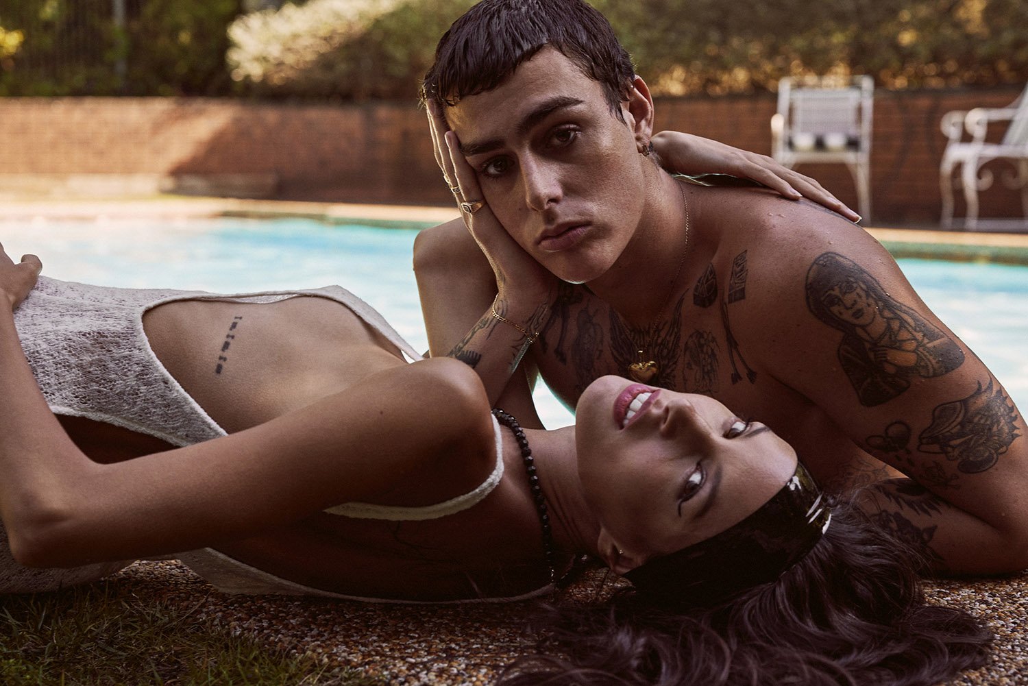 A young adult couple with tattoos lying next to a pool, with a brick wall and greenery in the background, captured in a medium shot.