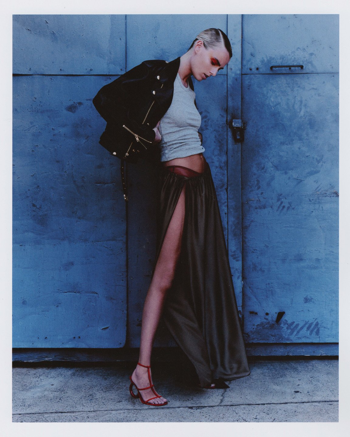A slender woman poses in front of a blue metal door, wearing a black jacket, grey top, a long slit skirt, and red strappy heels for a fashion shot.