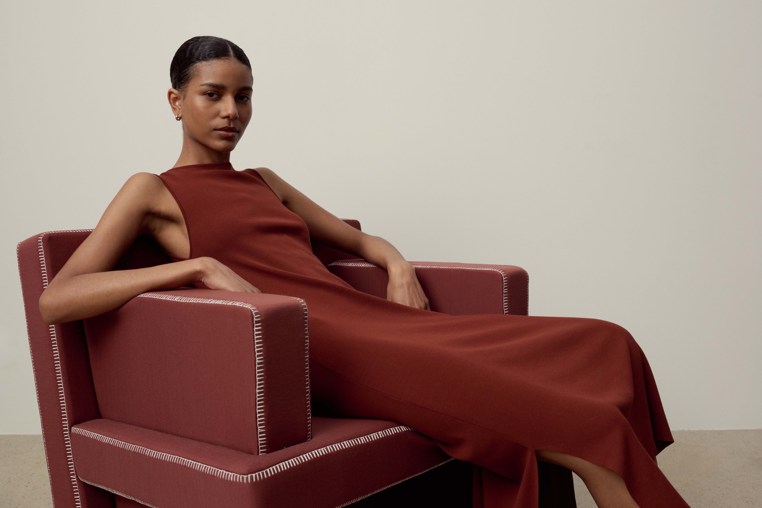 Elegant woman in a brown sleeveless dress sits gracefully in a matching chair against a neutral backdrop, showcasing fashion and style.
