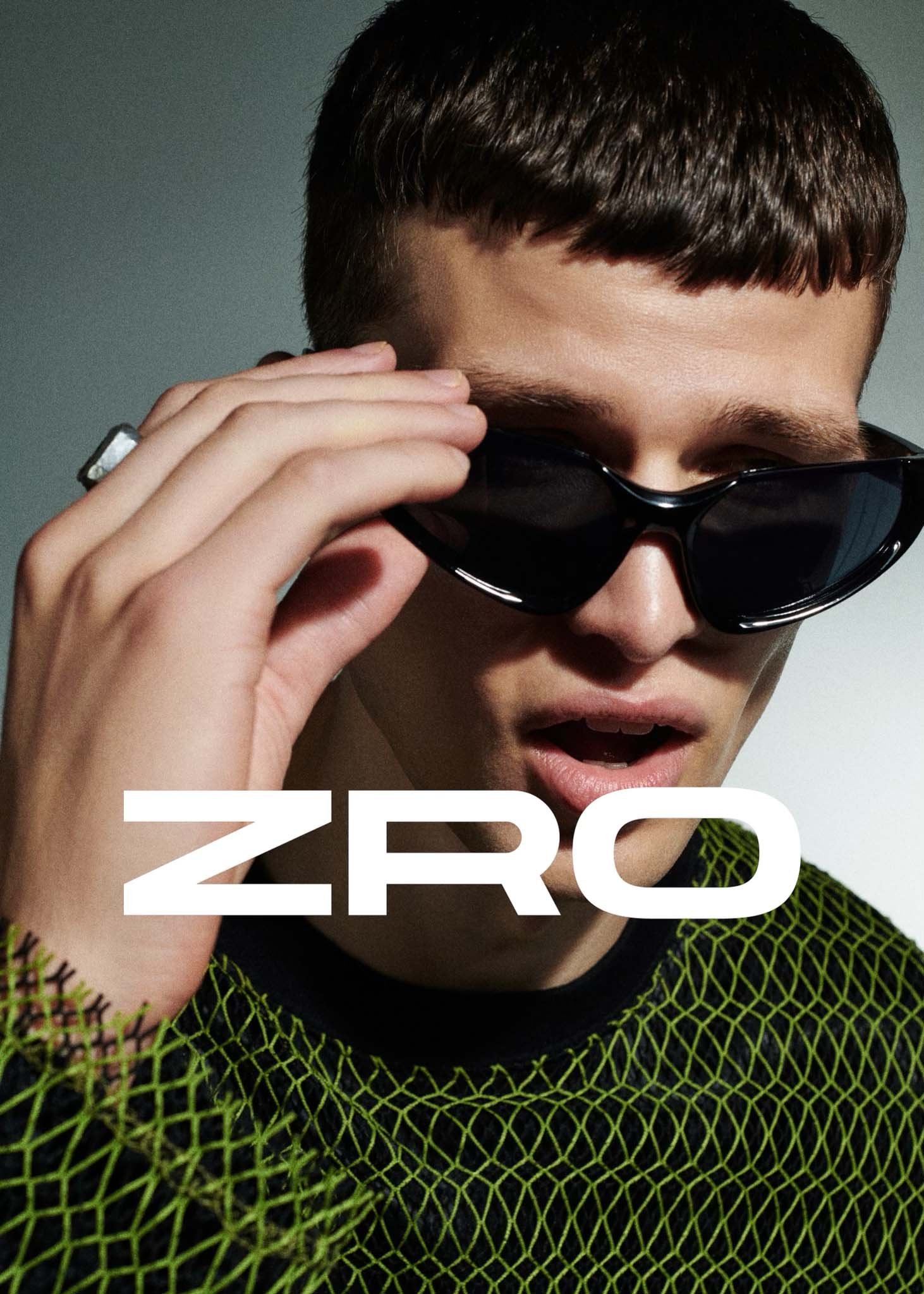 Close-up of a young man with dark hair wearing sunglasses and a green and black mesh shirt looking at the camera indoors.