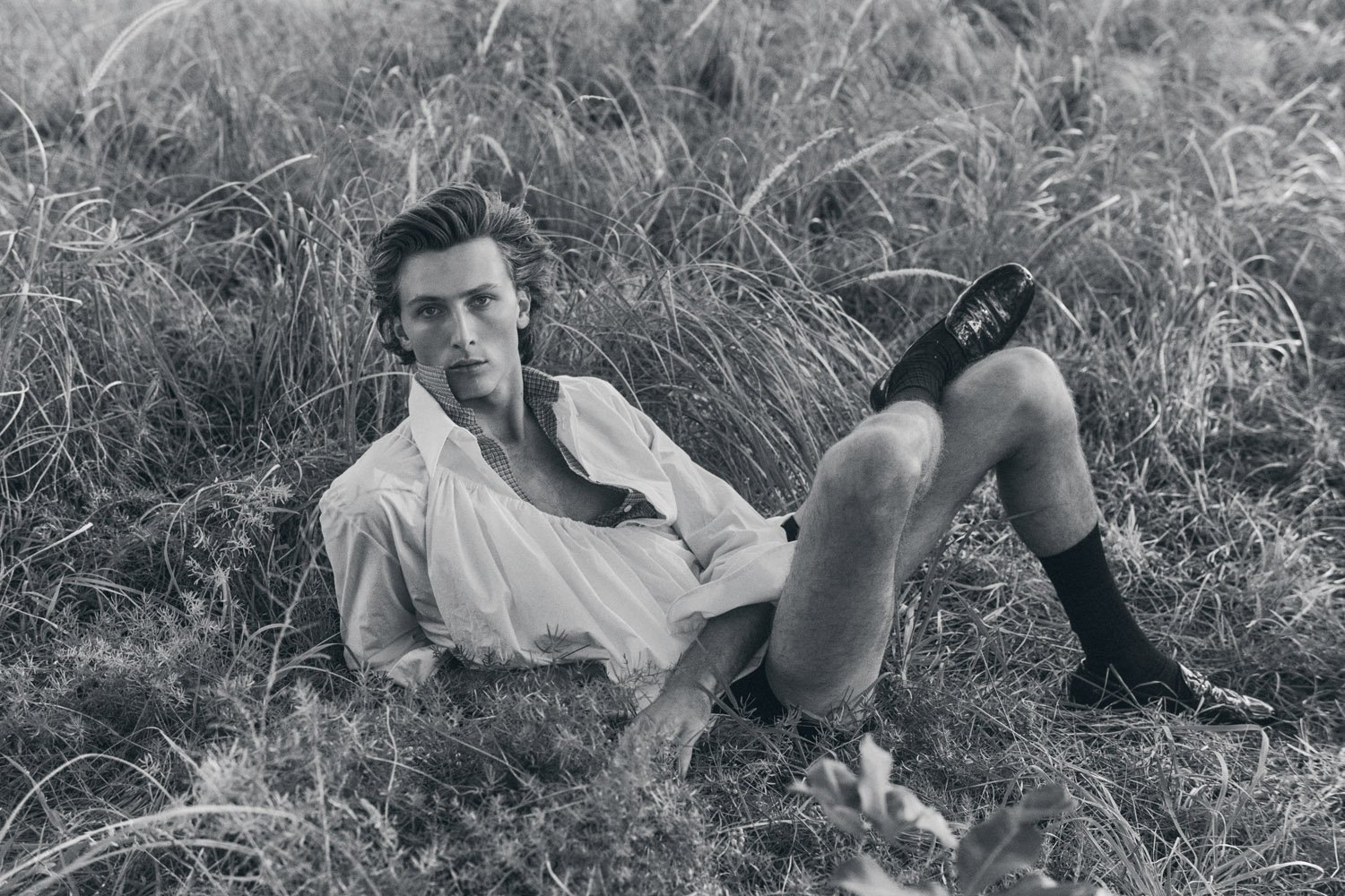 Monochrome portrait of a male model lying in tall grass, wearing an unbuttoned white shirt, dress shoes, and dark socks in a field.