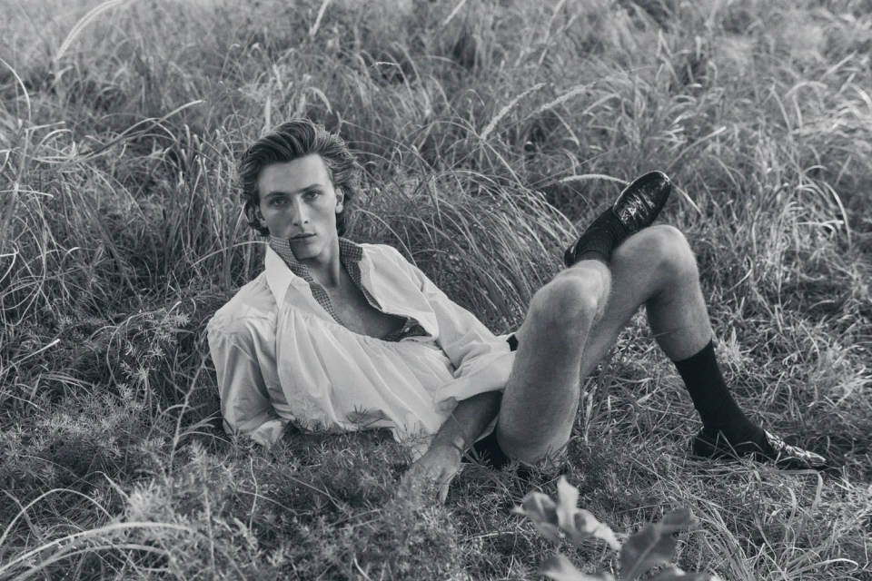 Monochrome portrait of a male model lying in tall grass, wearing an unbuttoned white shirt, dress shoes, and dark socks in a field.