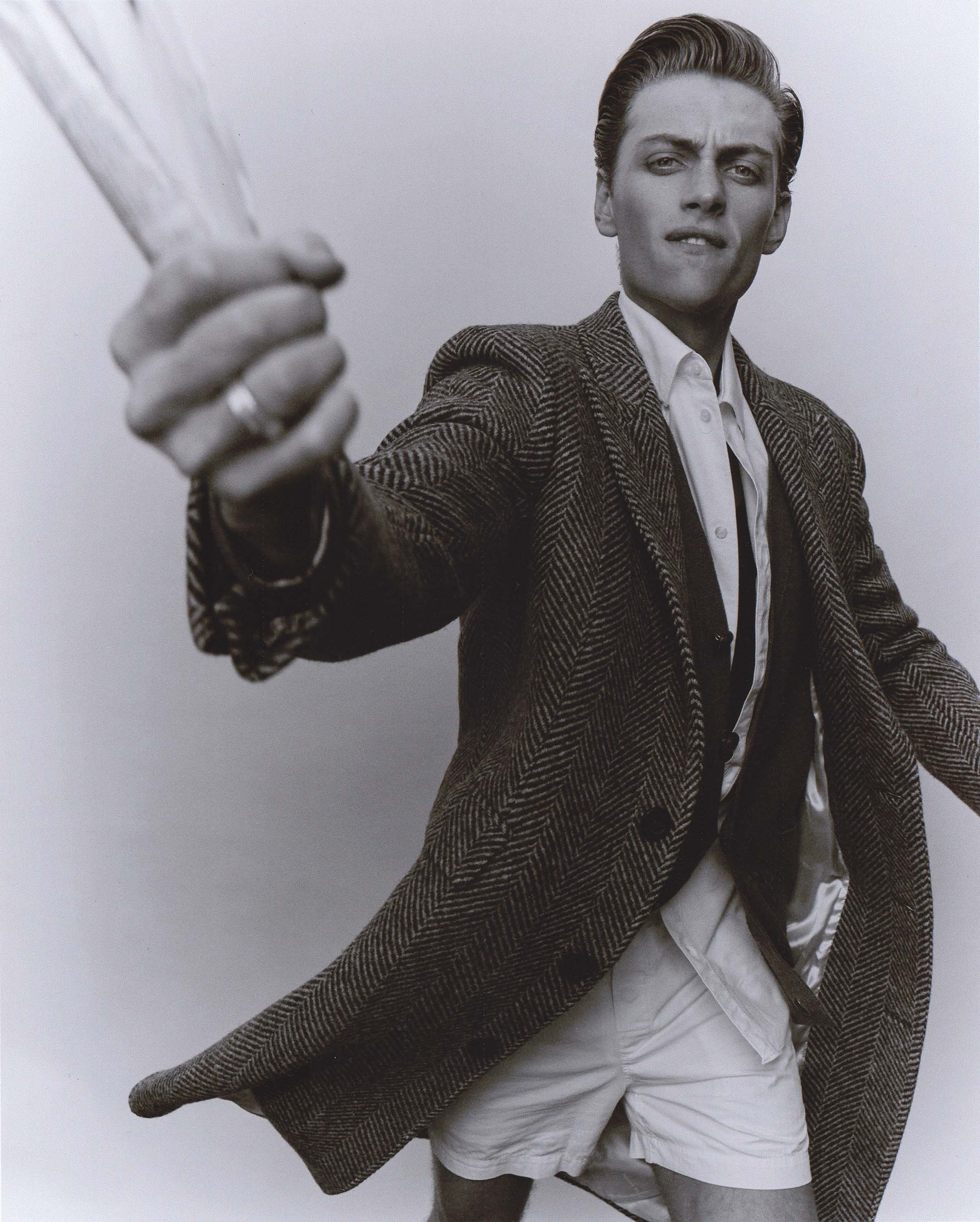 Monochrome portrait of a young adult male model wearing a herringbone coat, white shirt, and tie, holding up a bundle of newspapers.
