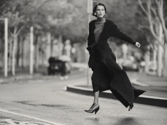 Black and white editorial fashion image of a woman in a long coat leaping across a wet city street, styled by Patrick Zaczkiewicz.