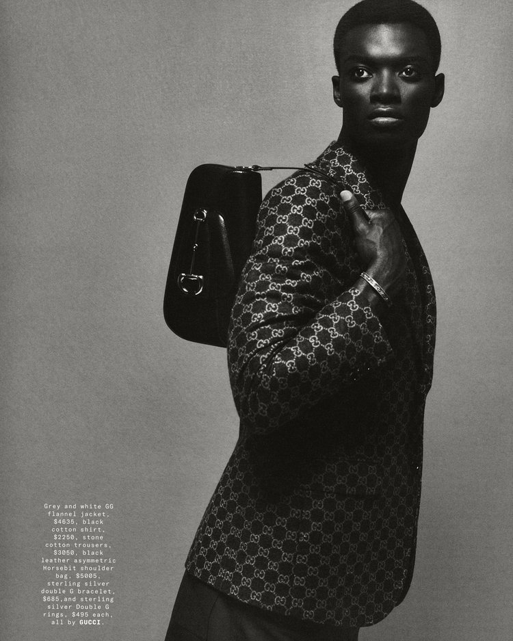 Fashion shot of an African American male model in a Gucci jacket with a black shoulder bag, posing against a grey backdrop.