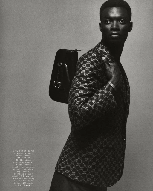 Fashion shot of an African American male model in a Gucci jacket with a black shoulder bag, posing against a grey backdrop.