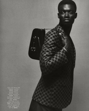 Fashion shot of an African American male model in a Gucci jacket with a black shoulder bag, posing against a grey backdrop.