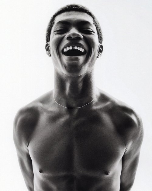 Monochrome portrait of an African-American man laughing with a missing front tooth, bare-chested with a thin silver necklace against a white backdrop.
