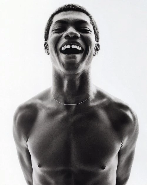 Monochrome portrait of an African-American man laughing with a missing front tooth, bare-chested with a thin silver necklace against a white backdrop.