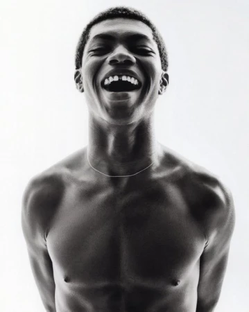 Monochrome portrait of an African-American man laughing with a missing front tooth, bare-chested with a thin silver necklace against a white backdrop.