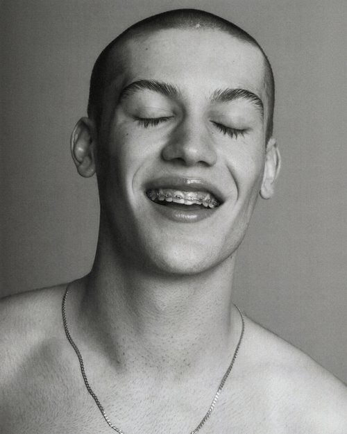 Monochrome portrait of a young adult male with closed eyes, smiling and wearing braces and a chain necklace against a neutral background.