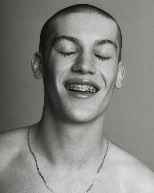 Monochrome portrait of a young adult male with closed eyes, smiling and wearing braces and a chain necklace against a neutral background.